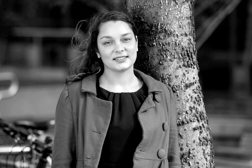 A woman with brown hair stands next a tree smiling.