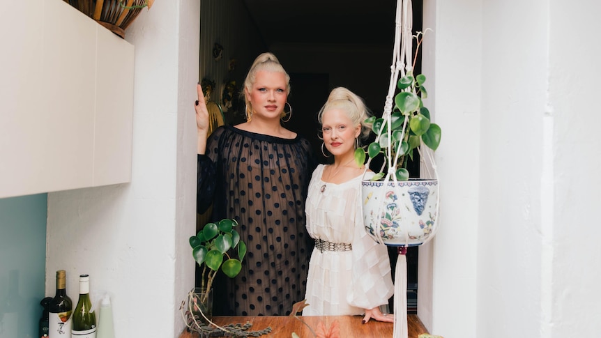 Two white women with matching platinum blonde hair stand in front of an open window. One wears black, the other wears white.