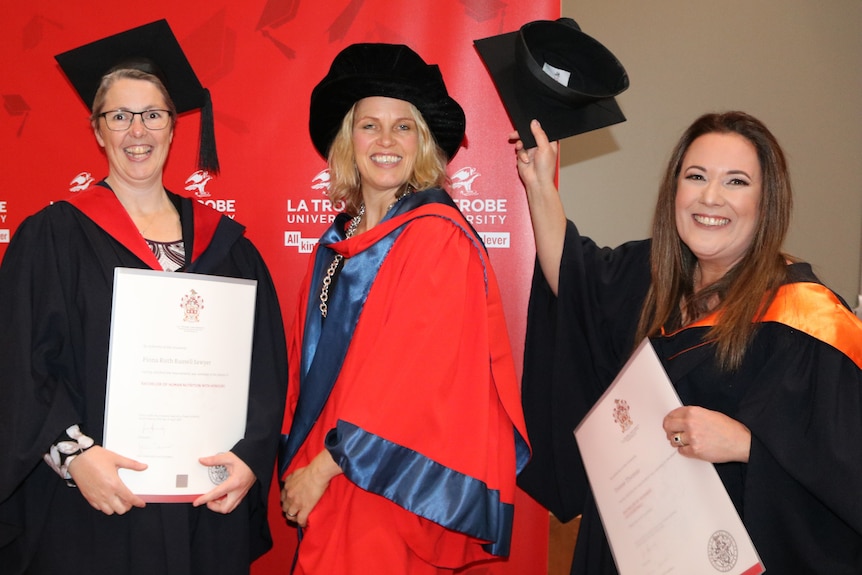 Three women smiling at a camera wearing university graduation gown and cap
