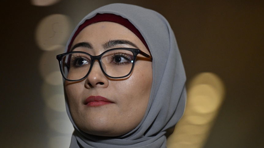 A young woman wearing thin glasses, light red lipstick and a grey hijab looks to her right in closeup