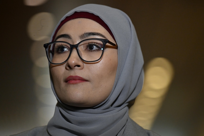 A young woman wearing thin glasses, light red lipstick and a grey hijab looks to her right in closeup