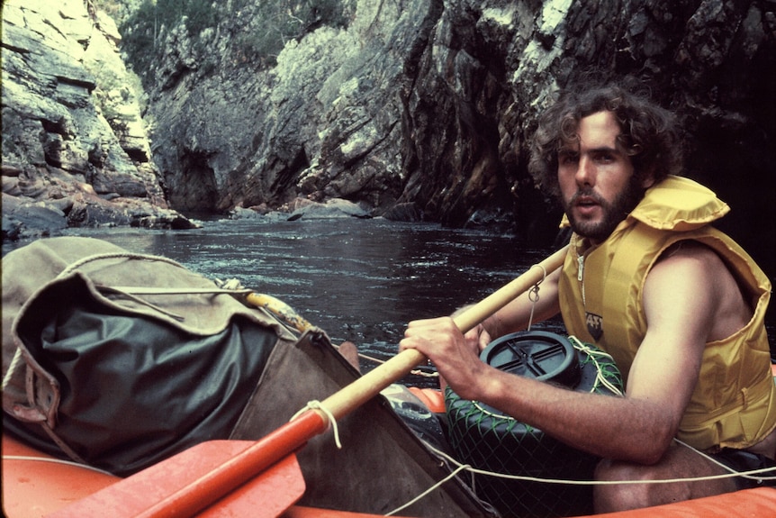 A man with shaggy hair and a beard sitting in a kayak holding a paddle.