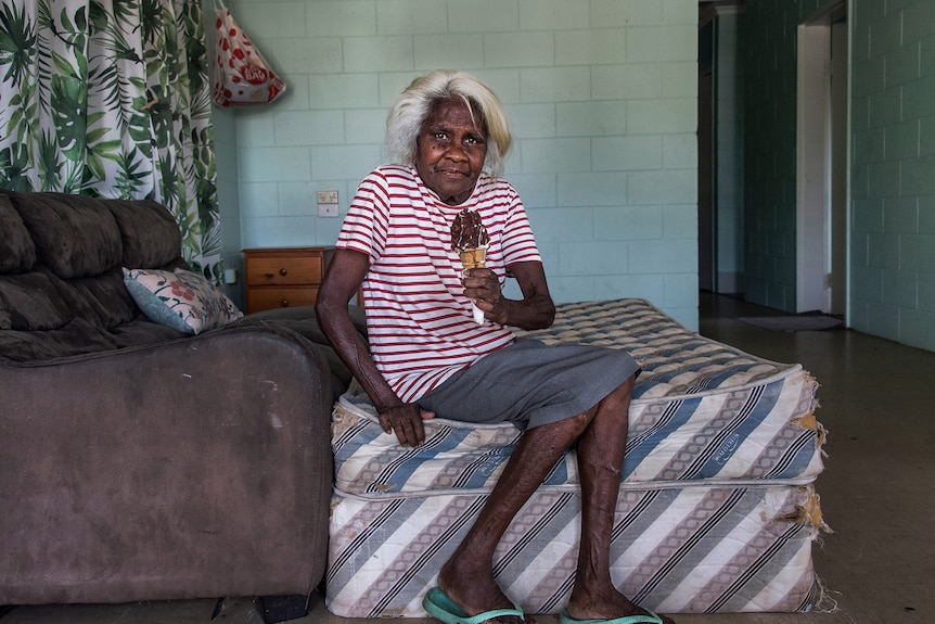 A woman sits on a bare mattress in a living room holding an ice cream.