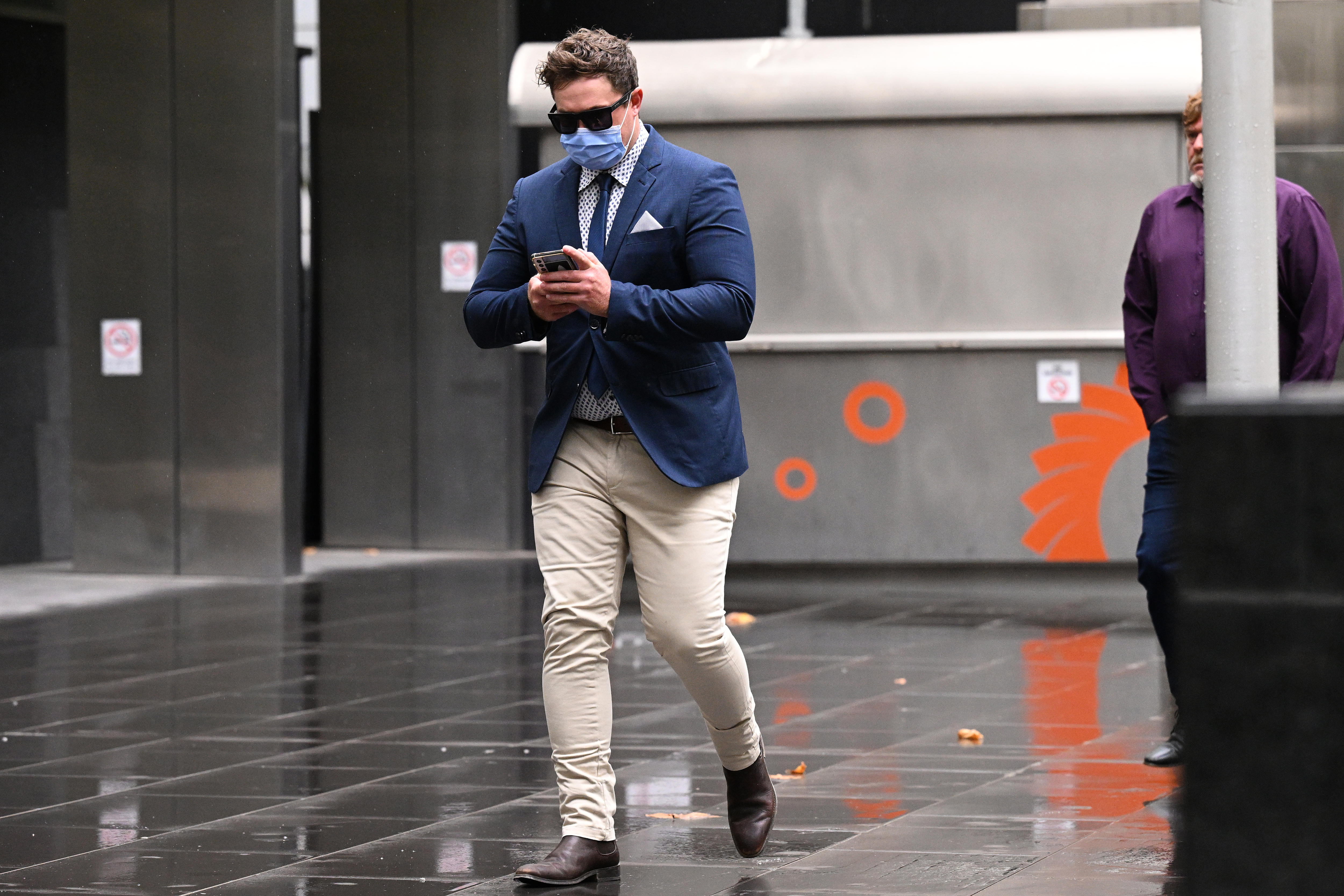 A man in moleskin pants and a jacket and tie, wearing a mask, walks on his phone, outside court.