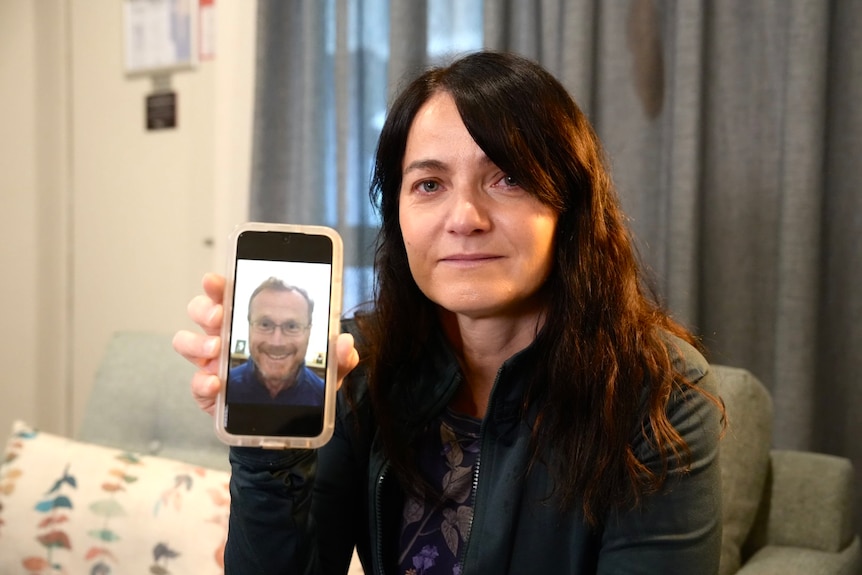 A woman holds up her smart phone with a photo of her partner smiling on it.