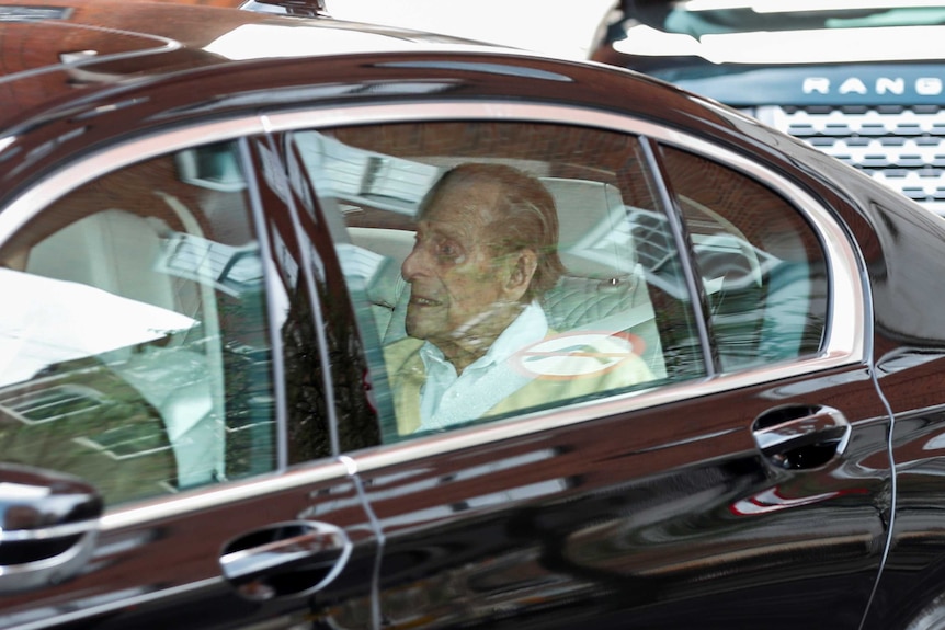 Prince Philip, wearing a bone cardigan, sits in the boot of a black car.