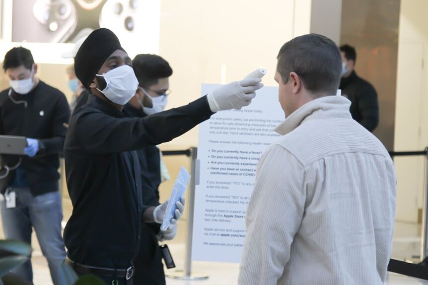 An employee wearing a face mask uses a digital thermometer to check the temperature of a man outside an Apple store