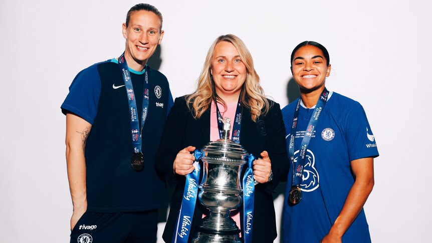 Chelsea WSL manager Emma Hayes holds the FA Cup while standing between players Ann-Katrin Berger and Jess Carter.