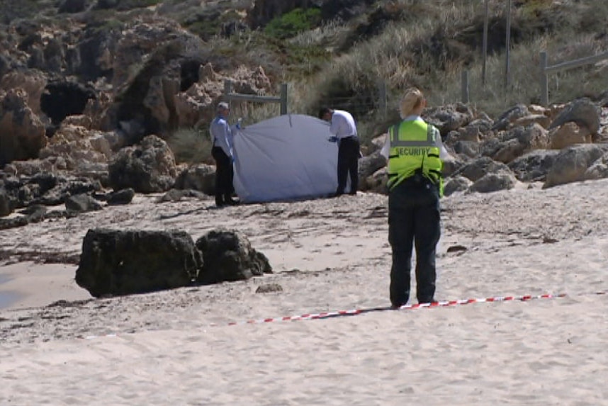 A security guard in a hi-vis vest stands in front of two police officers who are holding a white sheet up on a beach.