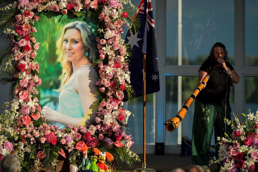 A photo of Justine Damond Ruszczyk is seen with flowers around it and a lady playing a didgeridoo in the background.