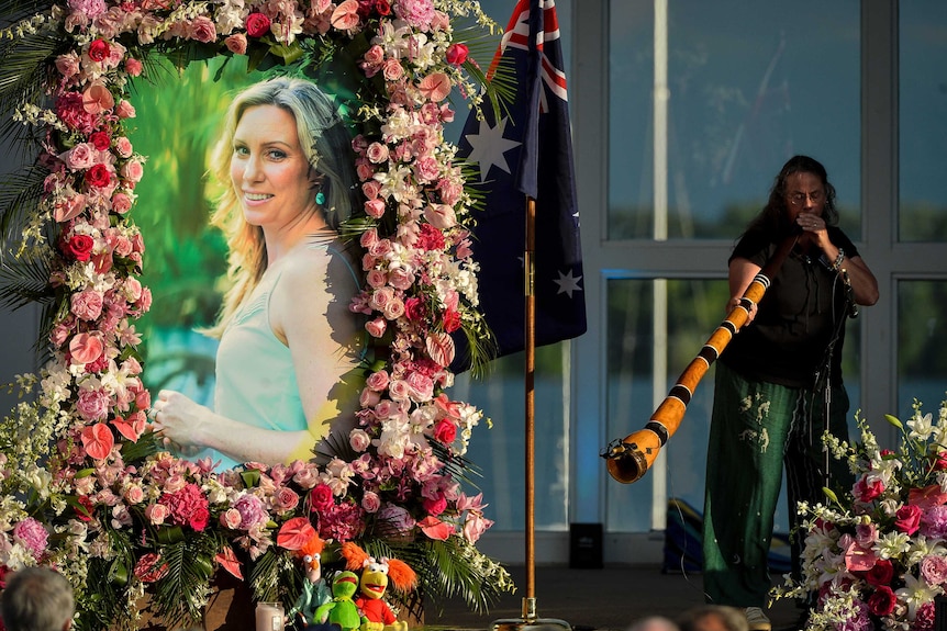A photo of Justine Damond Ruszczyk is seen with flowers around it and a lady playing a didgeridoo in the background.