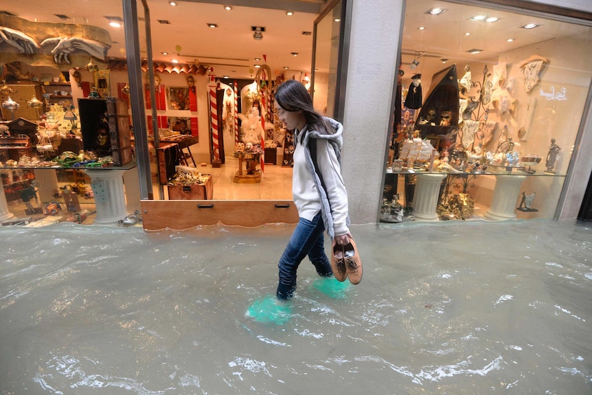 A woman walks through a flooded street in Venice, she is holding her shoes. The water is halfway up her shins.