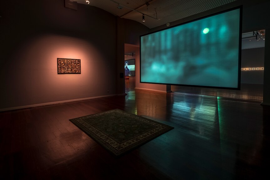 Darkened gallery with a persian rug on floor in front of a large video monitor with blurry image, and a painting on one wall.