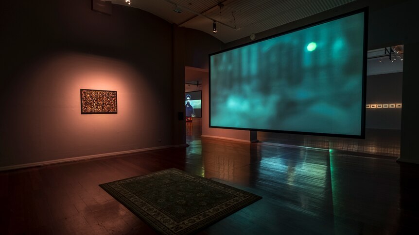 Darkened gallery with a persian rug on floor in front of a large video monitor with blurry image, and a painting on one wall.