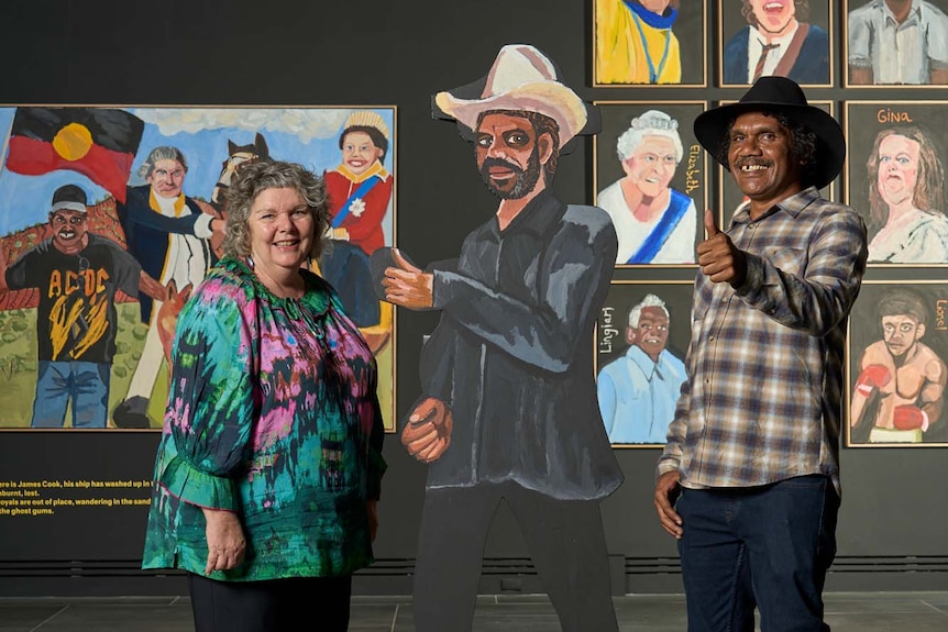 Vincent Namatjira gives the thums up, standing next to a prize-winning portrait of him also giving a thumbs 
