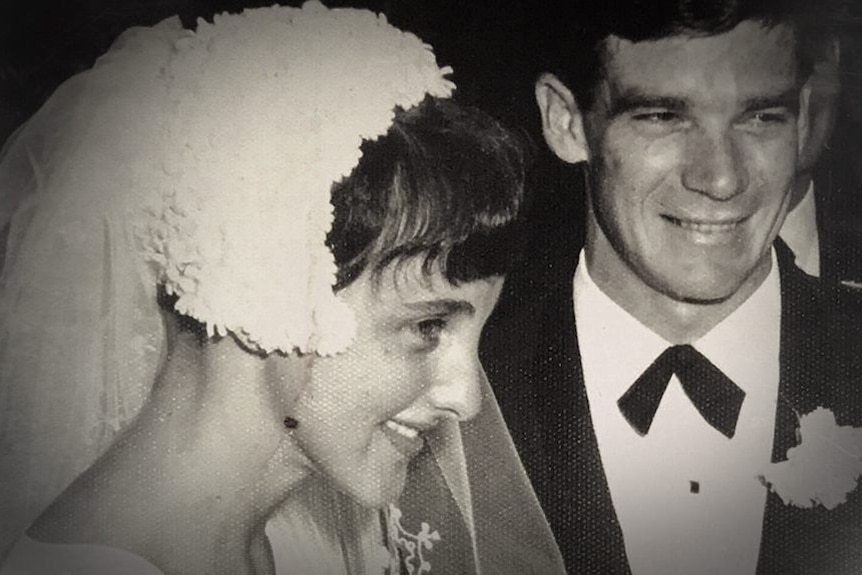 Marion Barter wearing a white wedding dress, heavy head dress, and Johnny Warren in a suit, black tie, flower, getting married.