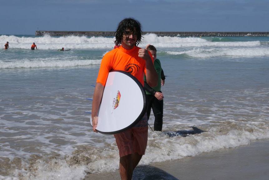 A teenager walks out from ocean carrying surf board and gives thumbs up