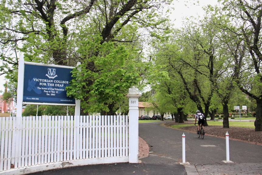 A blue and white sign stands beside a tree-lined drive way to a school for deaf students.