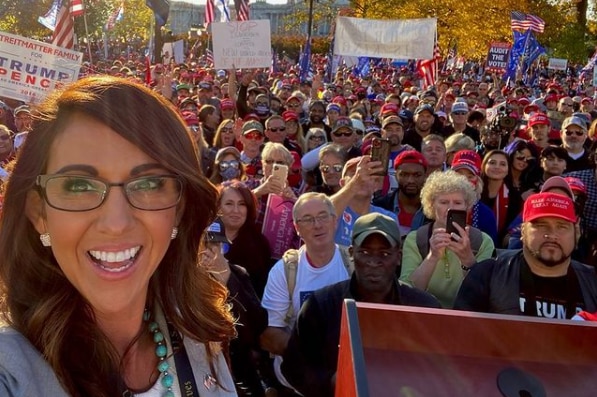 A woman with brown hair and waring glasses and earrings smiles in front of a crowd of people holding Trump signs.