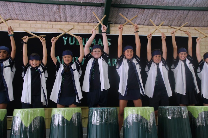 Yuluma Primary School students stand on stage in a row holding drum sticks above their heads.