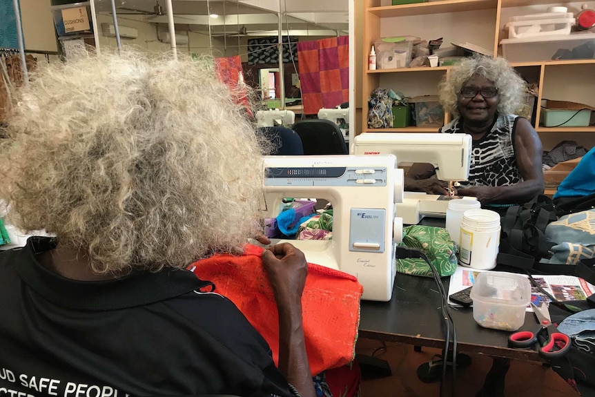 Two Aboriginal women sit in either side of a table and use sewing machines.