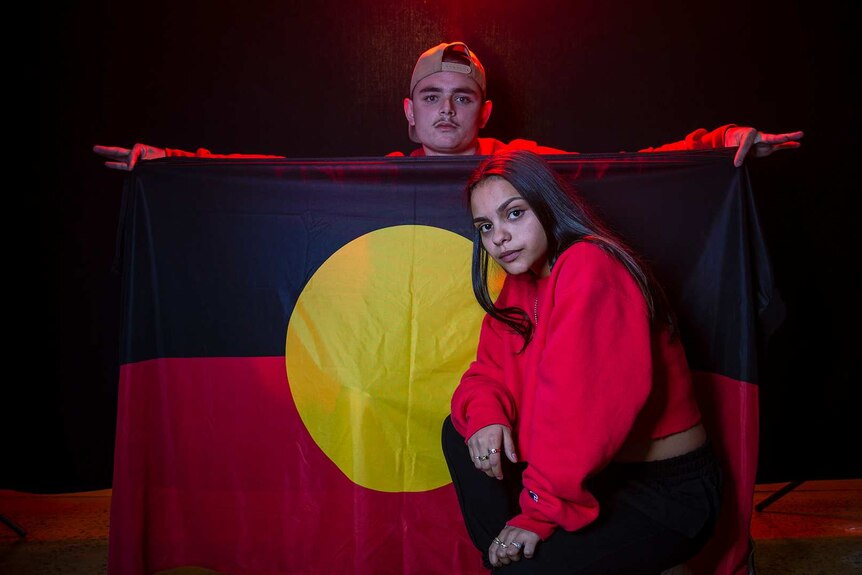 Colour photo of Braiden Unwin holding Aboriginal flag and Lakesha Grant, in front of black background with some red light.