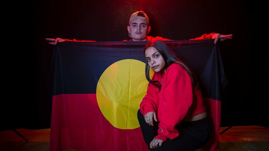 Colour photo of Braiden Unwin holding Aboriginal flag and Lakesha Grant, in front of black background with some red light.
