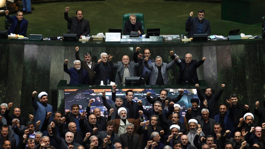 A crowd of Iranian MPs, all men, throw their fists into the air and are pictured mid-shout on the Iranian parliament's floor.