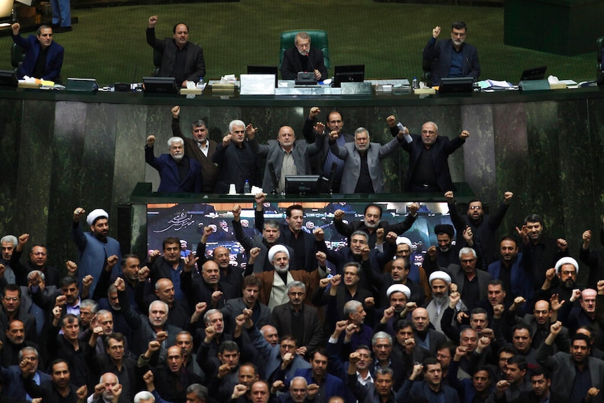 A crowd of Iranian MPs, all men, throw their fists into the air and are pictured mid-shout on the Iranian parliament's floor.