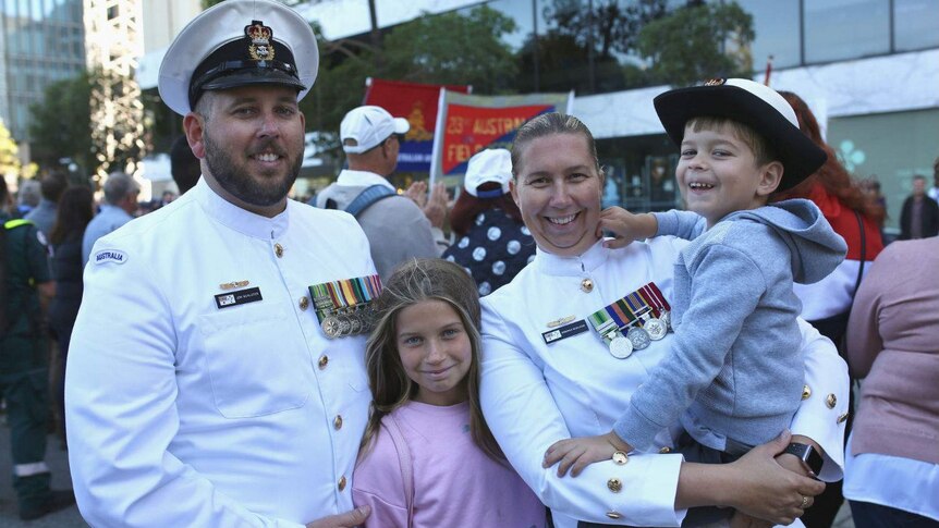Jon and Deborah Schluter wearing white navy attire on the street, with their two children, with the parade in the background.