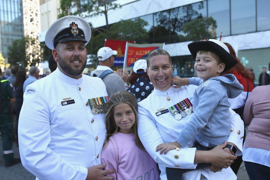 Jon and Deborah Schluter wearing white navy attire on the street, with their two children, with the parade in the background.