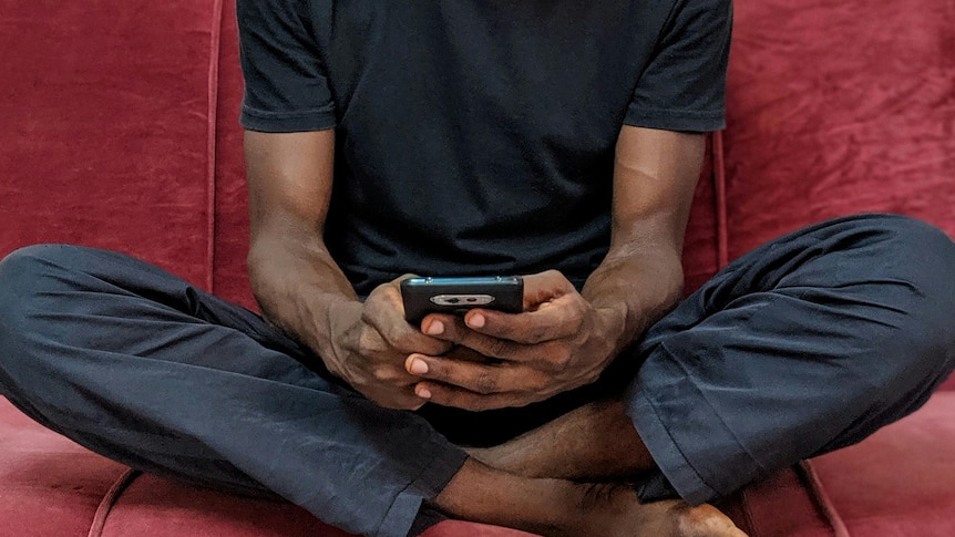 A man sits cross-legged on a red couch and looks down at his phone with a serious expression on his face.
