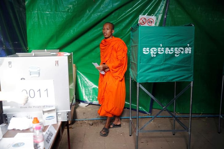 Monk dressed in orange robes walks toward ballot box holding his voting slip.