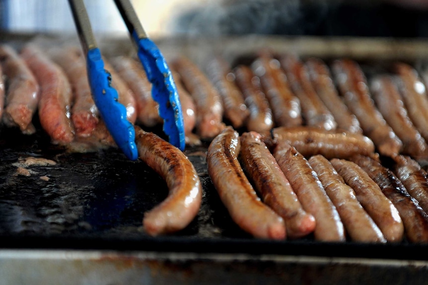 Sausages sizzling on a barbecue plate as a pair of tongs hover close by.