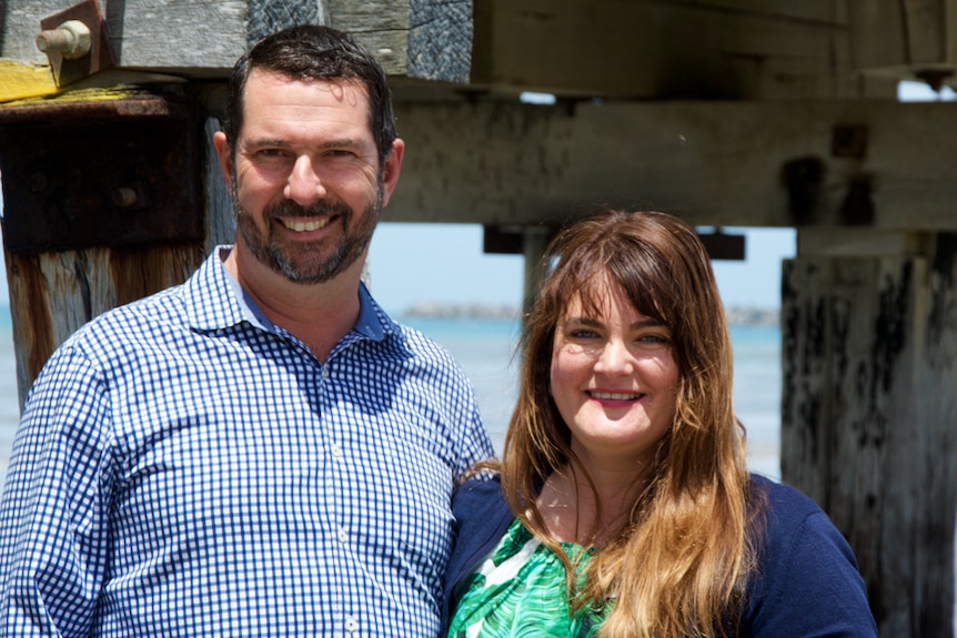 A man and a woman stand in front of wooden pylons and smile.