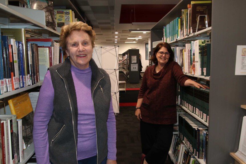 Two women stand between shelves of books.