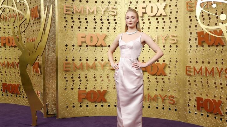 Sophie Turner puts her hands on her hips and poses against a gold background on the purple red carpet.