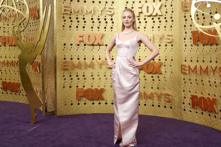 Sophie Turner puts her hands on her hips and poses against a gold background on the purple red carpet.