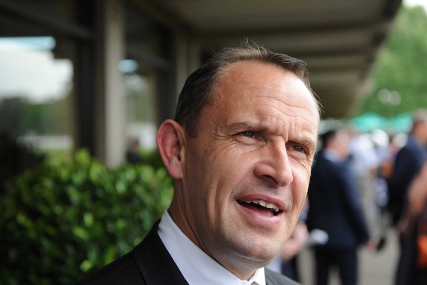 A racehorse trainer wearing a suit gives an interview in a racecourse mounting yard.