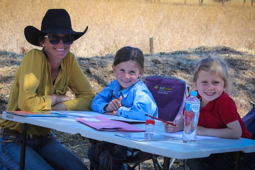 A mother sits at a camp table helping her two girls do school work