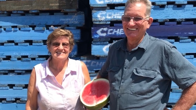 A couple stand in front of pallets holding a watermelon