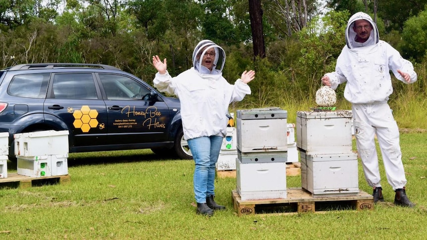 A man and woman in white bee suits hold a feeding tub over four boxes of bee hives