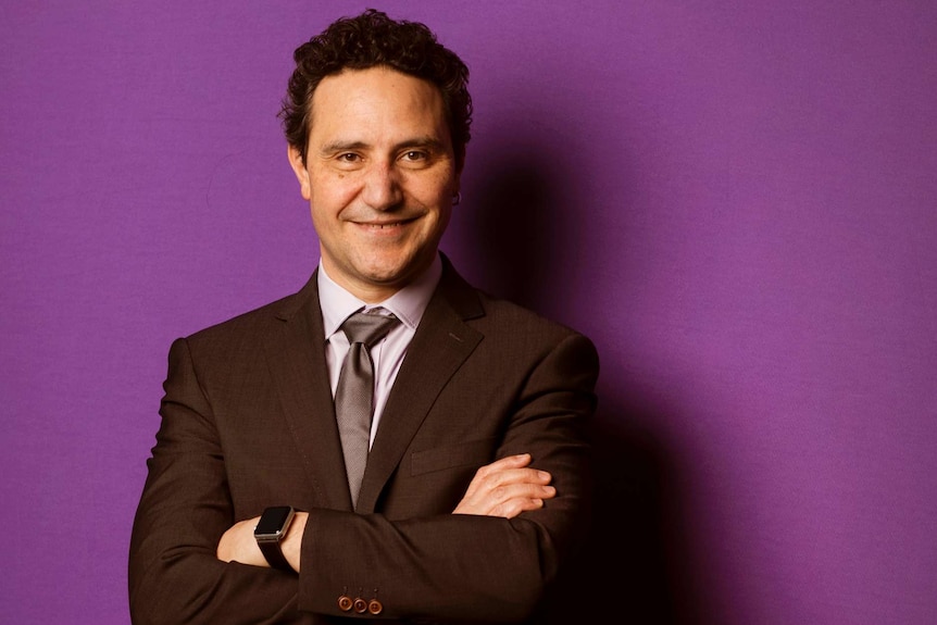 A man wearing a suit stands with his arms crossed in front of a purple wall