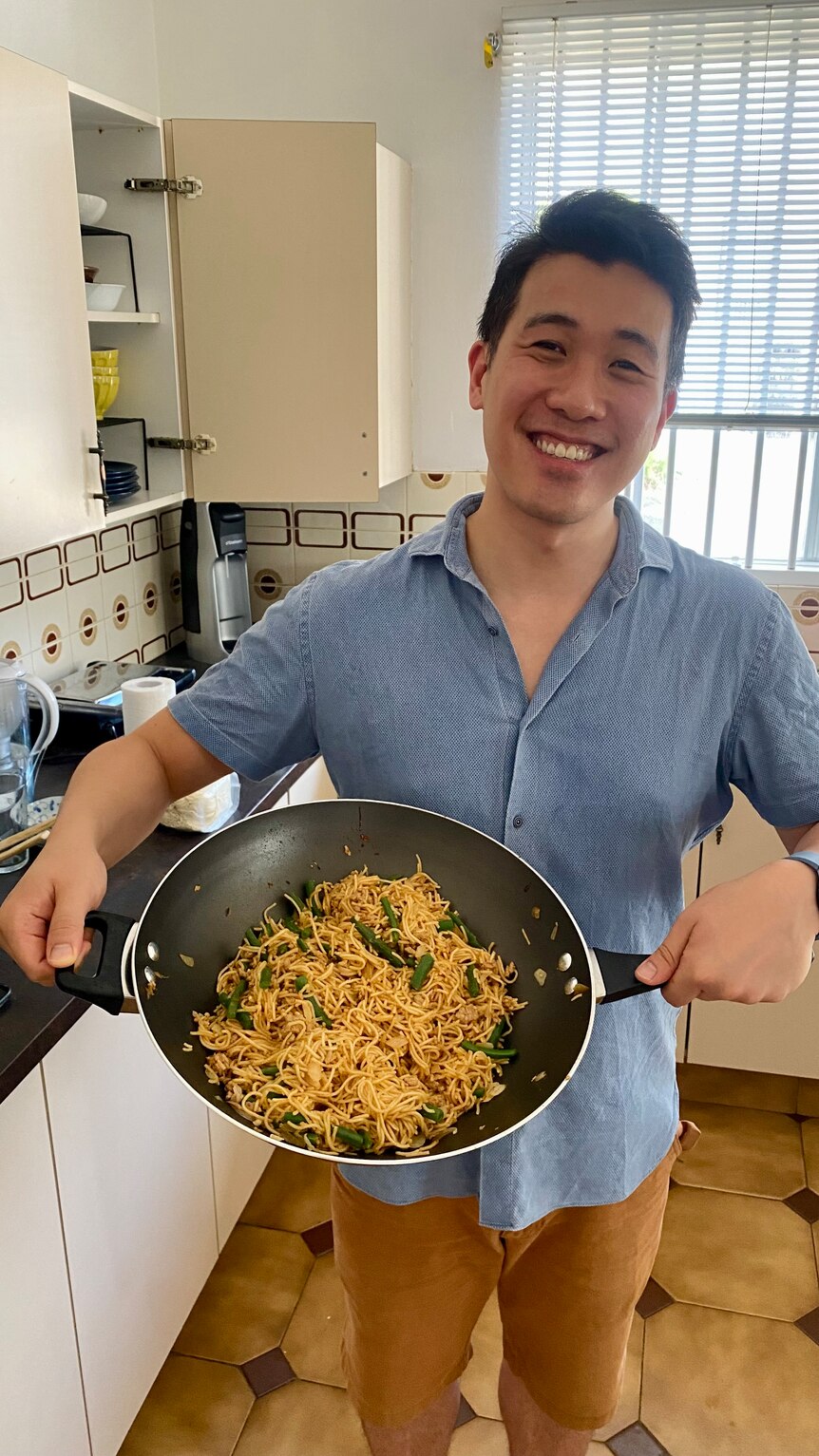 A young man of Asian appearance stanbds in a kitchen, smiling, and holding a wok filled with a noodle stir-fry.
