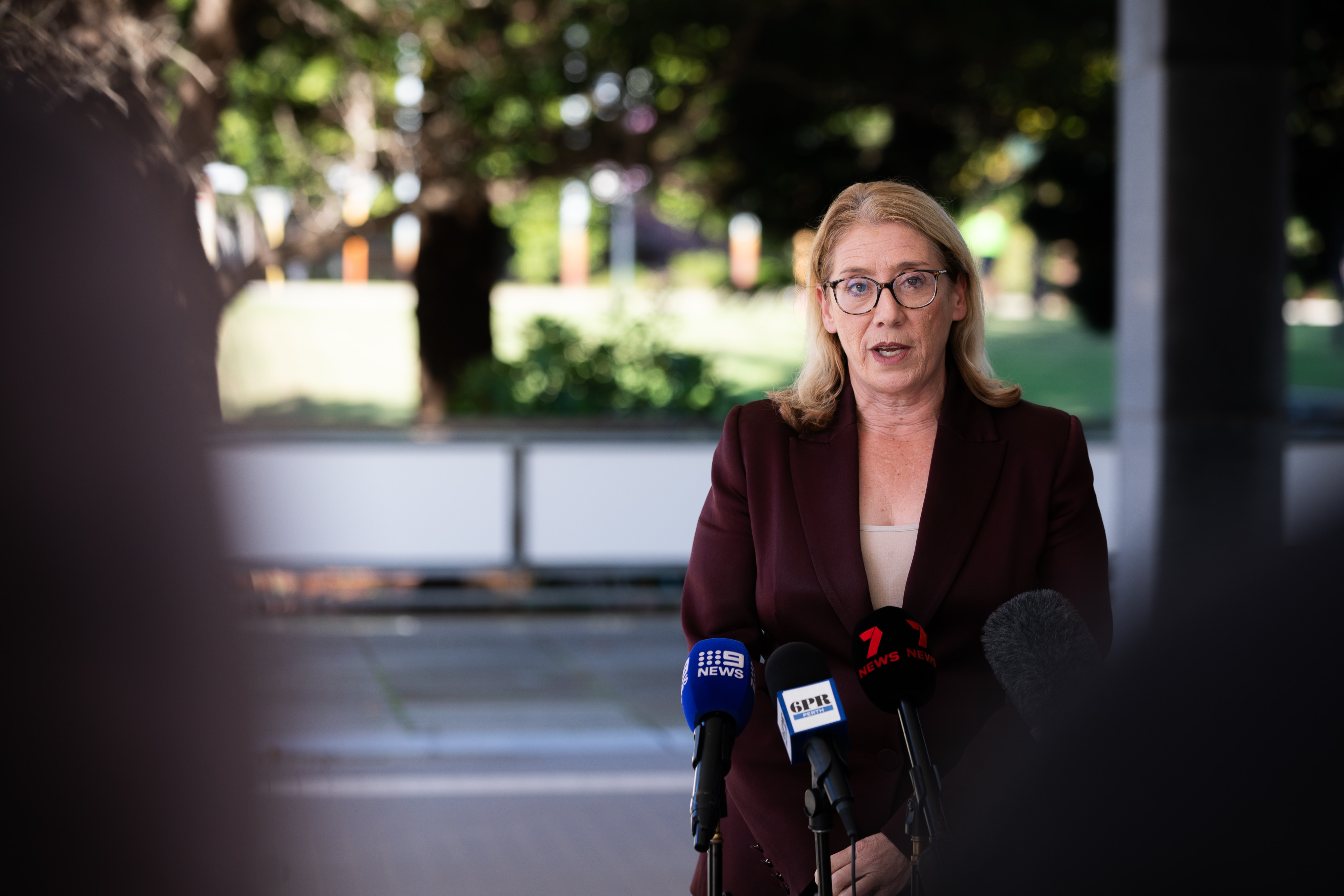 A woman with shoulder-length blonde hair, wearing a suit jacket, outside speaking to reporters.
