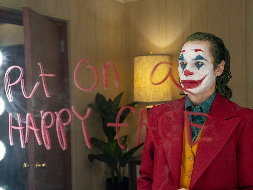Actor Joaquin Phoenix dressed as the Joker is looking into a mirror that has the words 'put on a happy face' written on it
