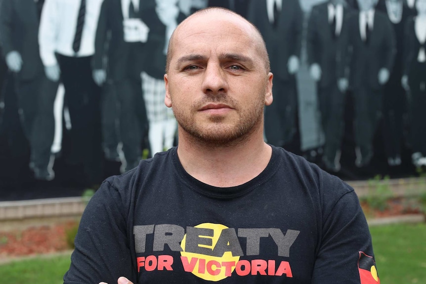 Jamie Lowe wears a Treaty t-shirt as he stands in front of a mural of past Victorian Aboriginal leaders.