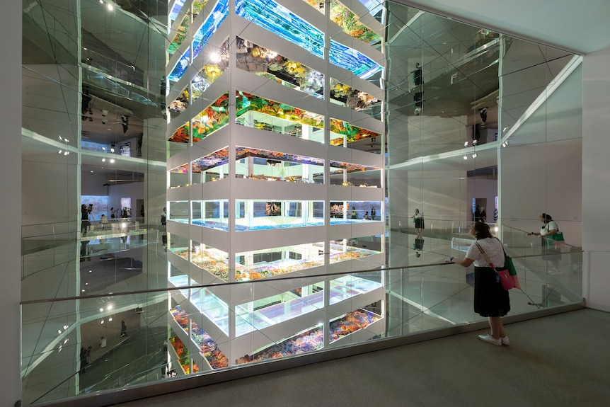 A large white structure of columns with flat platforms featuring illuminated colourful artworks towers in a gallery space.