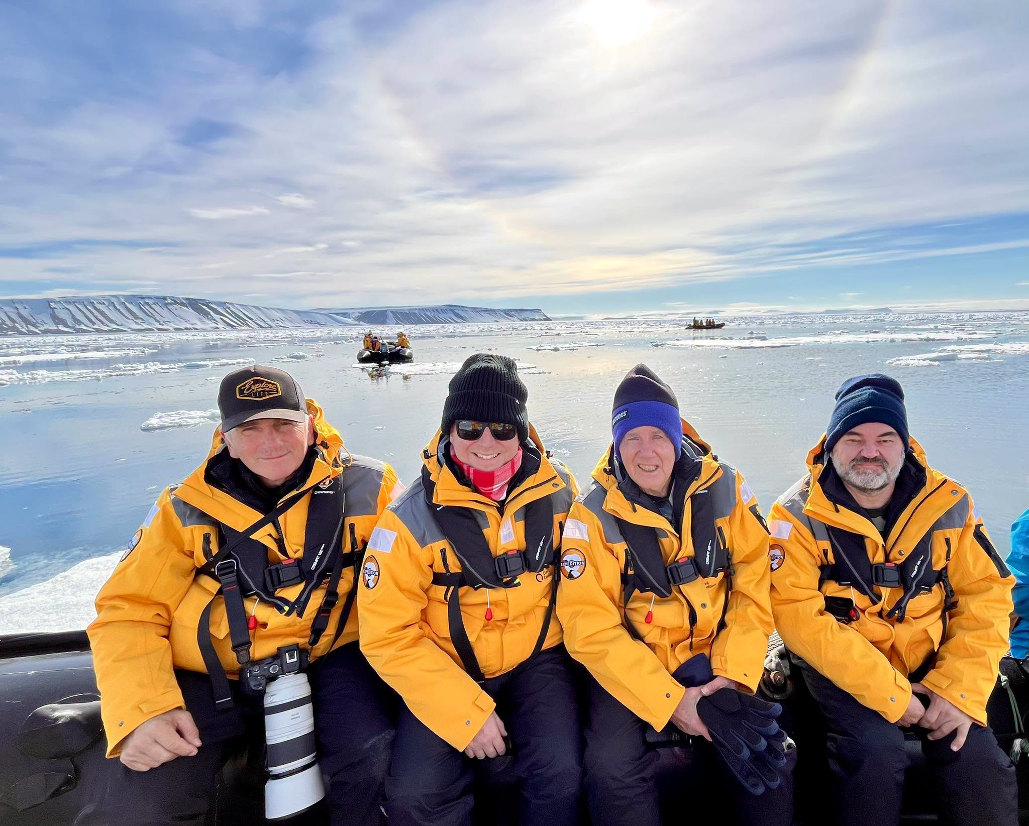 Four men in bright orange jackets and dark beanies sit in an inflatable boat in icy waters.