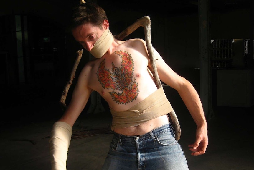 A man with bandage around his face and abdomen, with a skeleton in flames artwork visible on his chest.